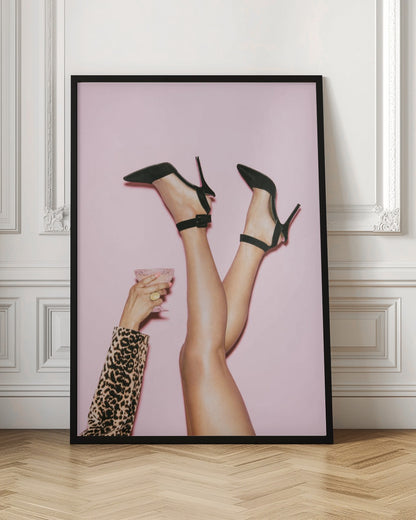 A woman's legs are kicked up in the air against a solid pink background. She is wearing black, pointed-toe, high-heeled shoes with ankle straps. A hand in a leopard print sleeve holds a pink, textured cocktail glass to the side. Artwork