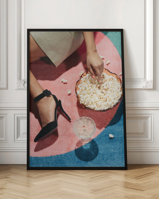 A high-angle photograph of a woman sitting on a vibrant pink and blue rug. She is wearing a chic black stiletto high heel and reaching into a glass bowl filled with popcorn. A crystal glass and scattered popcorn kernels are also on the rug. Artwork