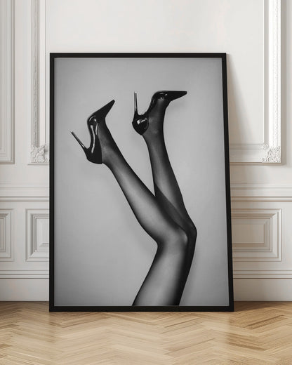 A black and white photograph of a woman's legs in the air, wearing sheer black stockings and shiny black stiletto high heels against a plain grey background. Poster