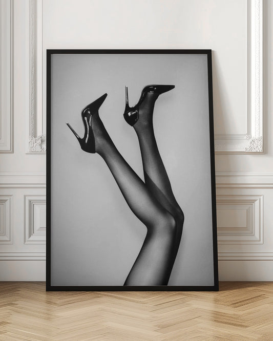 A black and white photograph of a woman's legs in the air, wearing sheer black stockings and shiny black stiletto high heels against a plain grey background. Poster
