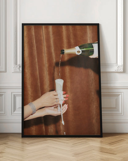 Champagne is poured from a bottle into a glass, overflowing with white foam that drips down onto two manicured hands with red nail polish holding it against a brown velvet curtain background. Poster
