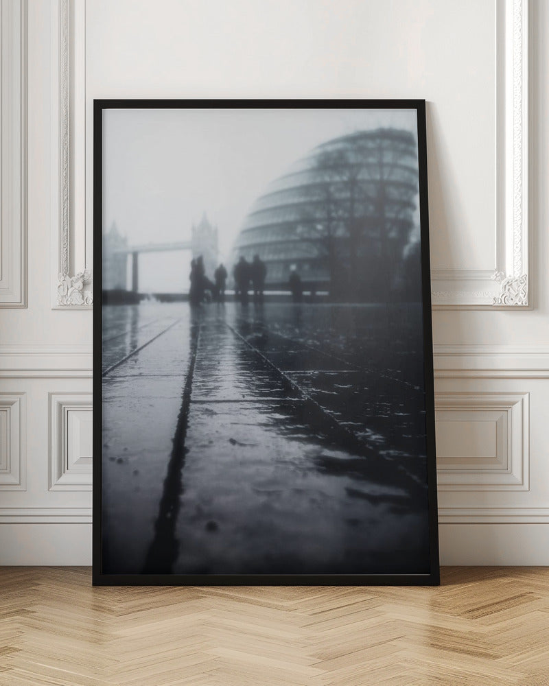 A moody, black and white, low-angle photograph of a wet city pavement in London. The foreground shows shimmering reflections on the dark, wet ground. In the blurry, foggy background, silhouettes of people stand near the modern City Hall building, with the iconic Tower Bridge visible further in the distance. Decor