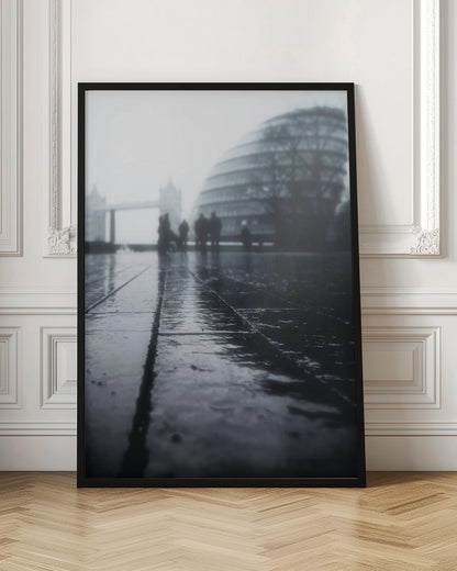 A moody, black and white, low-angle photograph of a wet city pavement in London. The foreground shows shimmering reflections on the dark, wet ground. In the blurry, foggy background, silhouettes of people stand near the modern City Hall building, with the iconic Tower Bridge visible further in the distance. Decor