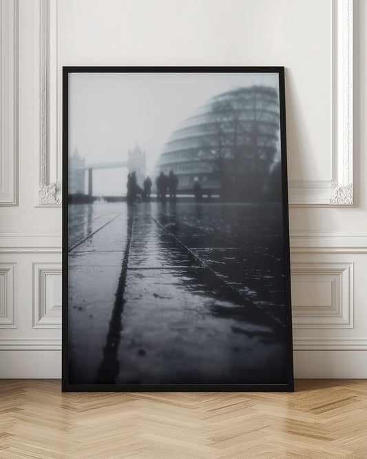A moody, black and white, low-angle photograph of a wet city pavement in London. The foreground shows shimmering reflections on the dark, wet ground. In the blurry, foggy background, silhouettes of people stand near the modern City Hall building, with the iconic Tower Bridge visible further in the distance. Decor