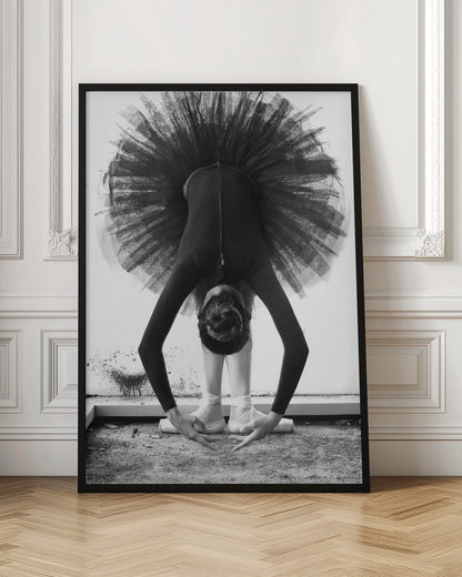A striking black and white photograph of a ballerina in a deep forward bend. Her body is clad in a black leotard and white tights, with a voluminous black tutu fanning out vertically behind her. Her hair is tied in a bun, and her hands gracefully reach towards her feet, which are in white pointe shoes. Artwork