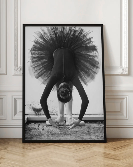 A striking black and white photograph of a ballerina in a deep forward bend. Her body is clad in a black leotard and white tights, with a voluminous black tutu fanning out vertically behind her. Her hair is tied in a bun, and her hands gracefully reach towards her feet, which are in white pointe shoes. Artwork