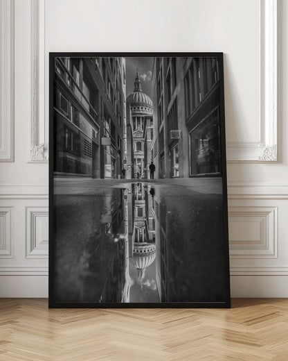 A dramatic black and white photograph taken from a low angle on a narrow city street, perfectly framing St. Paul's Cathedral in the distance. The foreground is dominated by a large puddle, which creates a stunning, clear reflection of the entire scene, including the cathedral, buildings, and sky. Print