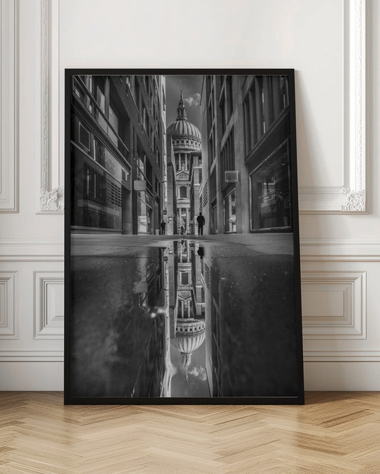 A dramatic black and white photograph taken from a low angle on a narrow city street, perfectly framing St. Paul's Cathedral in the distance. The foreground is dominated by a large puddle, which creates a stunning, clear reflection of the entire scene, including the cathedral, buildings, and sky. Print