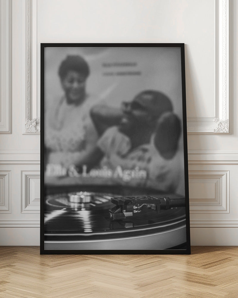 A black and white photograph focusing on a turntable's stylus resting on a spinning vinyl record. In the blurred background, the album cover for 'Ella & Louis Again' featuring Ella Fitzgerald and Louis Armstrong is visible. Poster