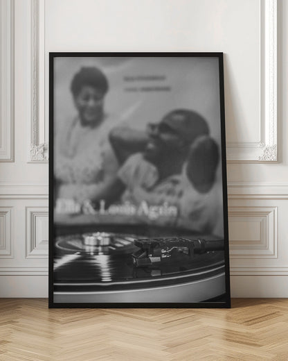 A black and white photograph focusing on a turntable's stylus resting on a spinning vinyl record. In the blurred background, the album cover for 'Ella & Louis Again' featuring Ella Fitzgerald and Louis Armstrong is visible. Poster