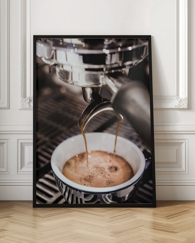 A close-up photograph of a shiny chrome espresso machine brewing a fresh shot of espresso. Two streams of rich, dark coffee pour from the portafilter into a white ceramic cup with a blue patterned rim, creating a light brown crema on top. Print