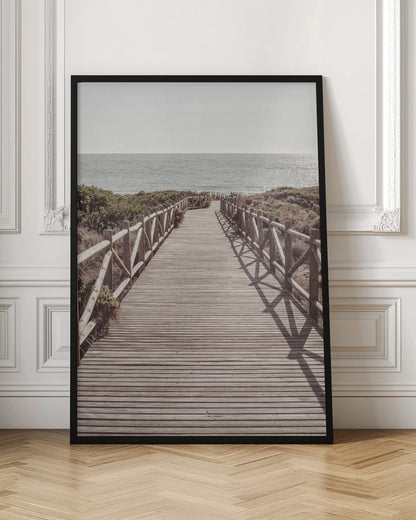 A long wooden boardwalk with rustic log railings leads the viewer's eye towards a calm, sunlit ocean under a hazy sky. Strong shadows from the railings fall across the path, creating a striped pattern. Poster