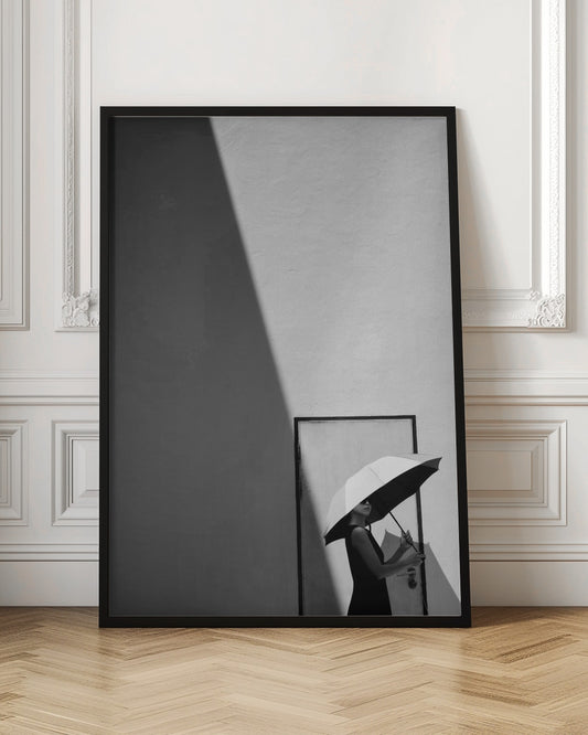 A minimalist black and white photograph of a woman in a black dress holding a two-toned umbrella. She stands in front of a door against a light wall, with a sharp, diagonal shadow dramatically cutting across the upper left of the image, creating a high-contrast, geometric composition. Decor