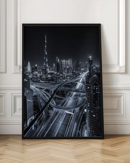 A cool-toned black and white photograph of the Dubai skyline at night. The iconic Burj Khalifa stands tall in the background, while the foreground is dominated by a massive, complex highway interchange with light trails from moving cars, creating a sense of motion and modernity. Poster