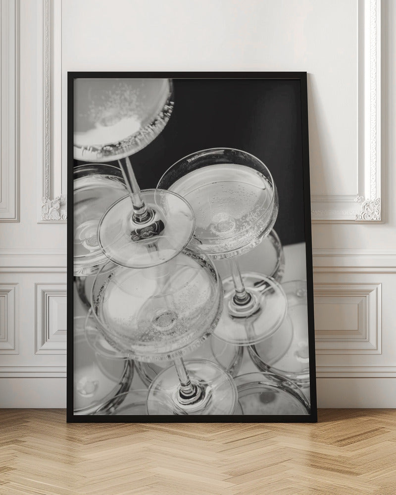 A close-up, high-angle photograph in black and white of a champagne tower. Several coupe-style glasses are stacked, filled with bubbly champagne that catches the light. The background is dark, creating a strong contrast and highlighting the elegant glassware. Decor