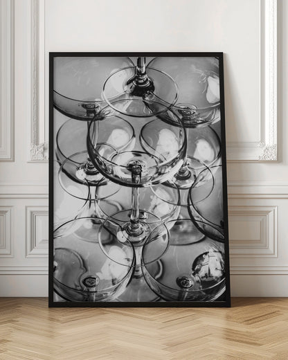 A black and white, overhead photograph of a tightly packed stack of empty champagne coupe glasses. The overlapping rims, stems, and bowls create a complex abstract pattern of circles and reflections, all enclosed within a simple black frame. Poster