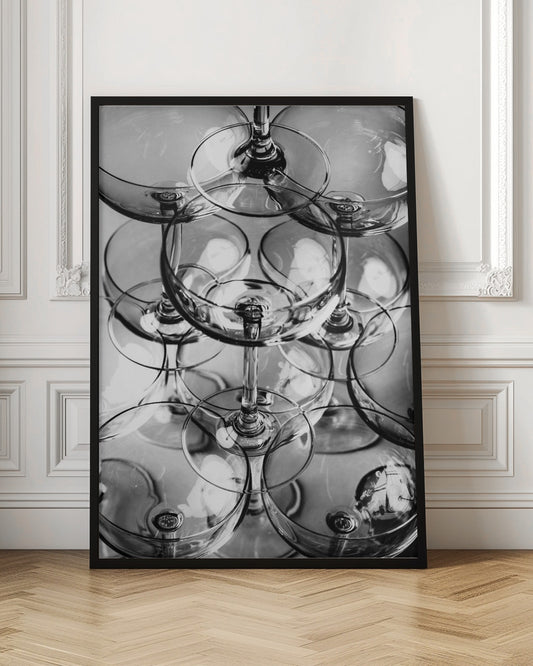 A black and white, overhead photograph of a tightly packed stack of empty champagne coupe glasses. The overlapping rims, stems, and bowls create a complex abstract pattern of circles and reflections, all enclosed within a simple black frame. Poster