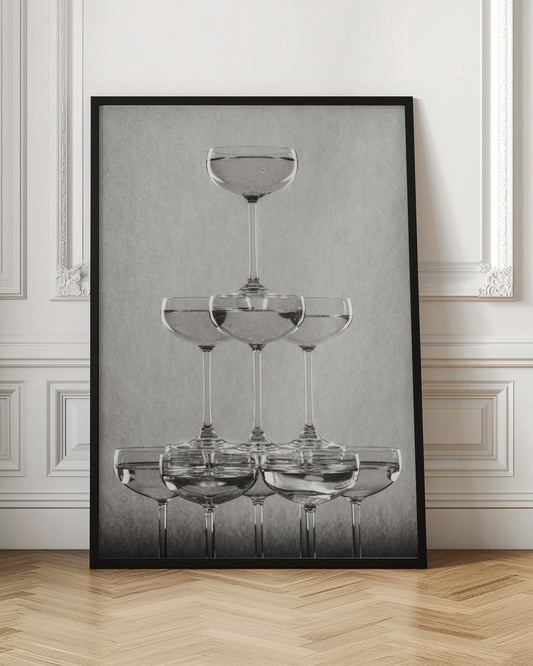A classic black and white photograph of a champagne tower, with coupe glasses stacked in a pyramid. The glasses are filled with bubbly liquid, and the composition is set against a textured light gray background and enclosed in a black frame. Poster