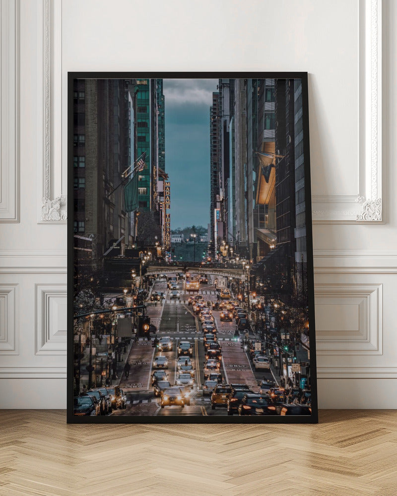A high-angle, vertical photograph looking down a bustling New York City street at dusk, framed by towering skyscrapers. The street below is packed with cars, their headlights creating a warm golden glow, contrasting with the cool, dark blue tones of the twilight sky and buildings. Poster