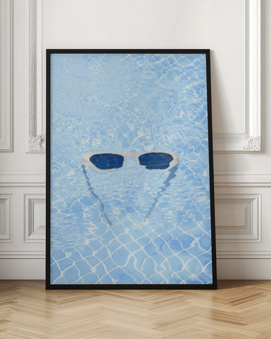 A pair of white-framed sunglasses with dark lenses floats on the surface of a bright blue swimming pool. The rippling water distorts the view of the tiled pool floor, and the sunglasses cast a faint shadow underneath. Print