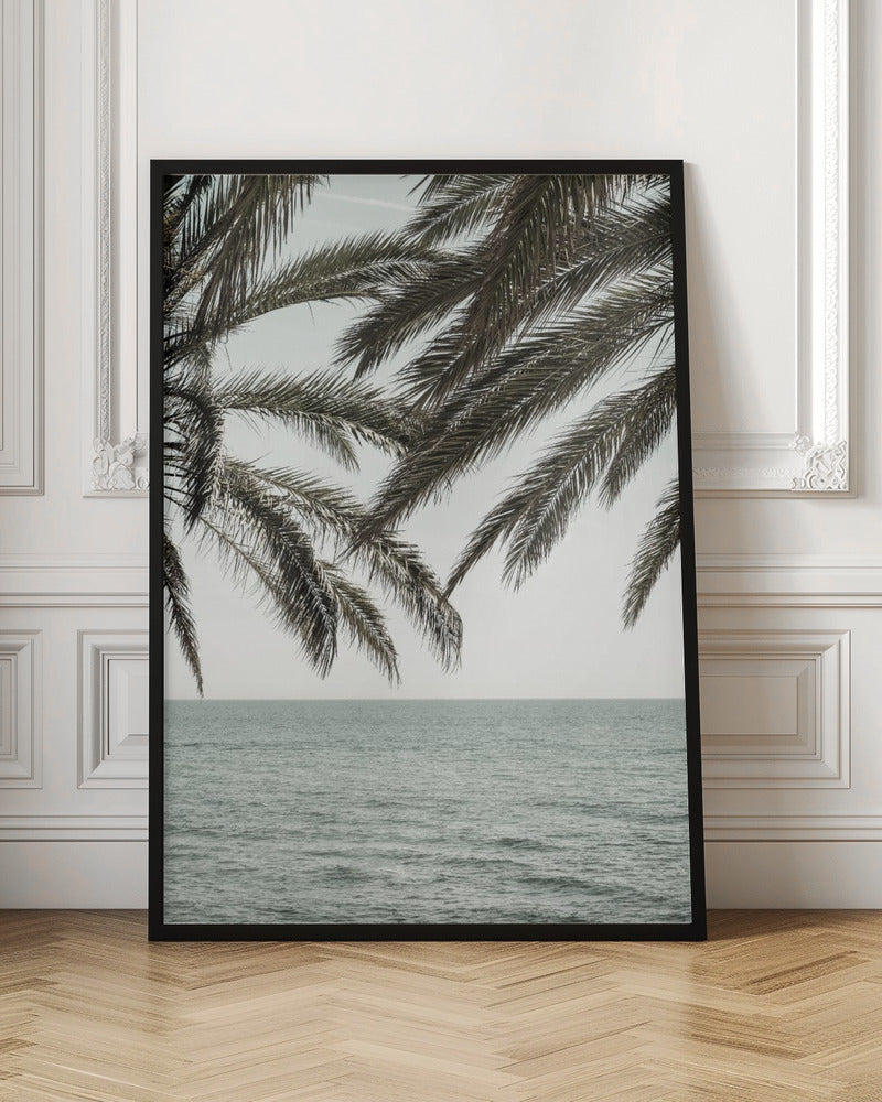 A close-up view of dark green palm tree fronds hanging over a calm, muted sage green ocean under a pale, overcast sky. The photograph has a serene and tranquil mood and is displayed in a black frame. Wall Art