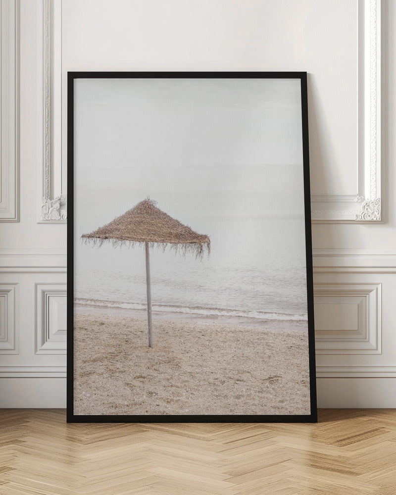 A minimalist photograph of a solitary thatched umbrella on a sandy beach, with calm, grey water and a hazy, overcast sky in the background, creating a serene and tranquil coastal scene. Poster