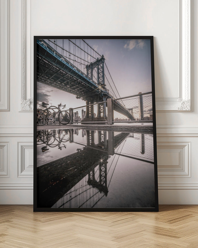 A dramatic low-angle photograph of the Manhattan Bridge, with its intricate steel structure and cables reflected in a large puddle on the pavement in the foreground. Two bicycles are parked near a fence on the left, with the New York City skyline visible in the distance under a softly lit, cloudy sky. Wall Art