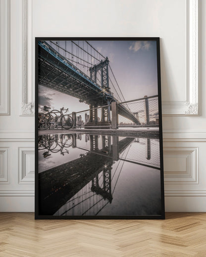 A dramatic low-angle photograph of the Manhattan Bridge, with its intricate steel structure and cables reflected in a large puddle on the pavement in the foreground. Two bicycles are parked near a fence on the left, with the New York City skyline visible in the distance under a softly lit, cloudy sky. Wall Art