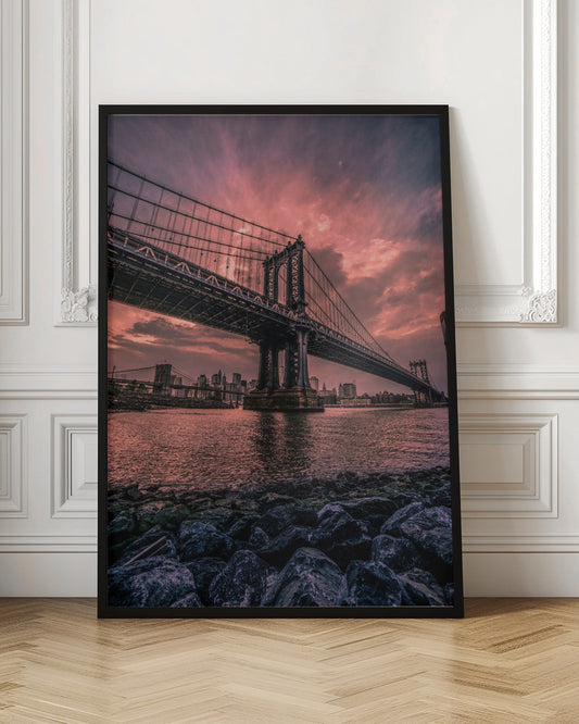 A dramatic low-angle view of the Manhattan Bridge over the East River at sunset. The sky is filled with pink and red clouds, which are reflected in the water below. The dark silhouette of the bridge contrasts with the vibrant sky, and the New York City skyline is visible in the background. In the foreground, dark, wet rocks line the riverbank. Decor
