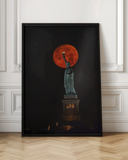 A striking night photograph of the Statue of Liberty perfectly silhouetted against a giant, vibrant red supermoon, which appears to be held aloft by the statue's torch against a deep black sky. Artwork