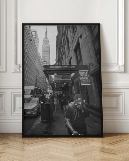A powerful black and white vertical photograph capturing a New York City street scene. An elderly Black man with a weary expression is in the foreground, walking along a crowded sidewalk. In the background, the Empire State Building towers over the cityscape, framed between buildings, with a sign for the Hotel Pennsylvania visible. Artwork