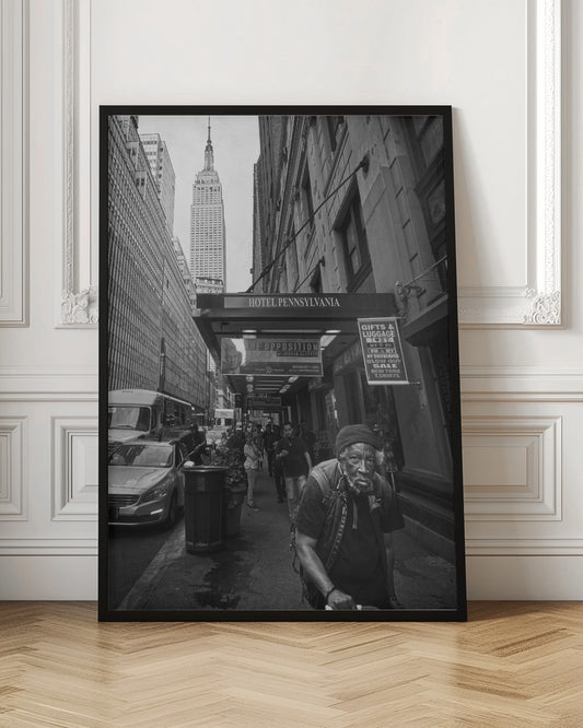 A powerful black and white vertical photograph capturing a New York City street scene. An elderly Black man with a weary expression is in the foreground, walking along a crowded sidewalk. In the background, the Empire State Building towers over the cityscape, framed between buildings, with a sign for the Hotel Pennsylvania visible. Artwork