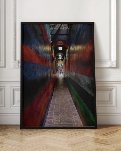 A long, narrow brick alleyway viewed from a one-point perspective, with walls painted in wide, colorful horizontal stripes of red, blue, and black, leading to a distant, sunlit exit where a person stands. Wall Art