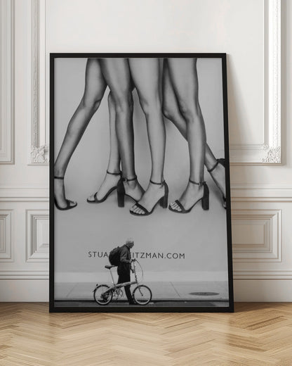 A black and white street photograph showing a stark contrast between a large fashion advertisement and a person on the sidewalk. The ad features multiple pairs of long female legs wearing high-heeled sandals. Below the ad, an older man with a backpack walks by, pushing a small foldable bicycle. Artwork
