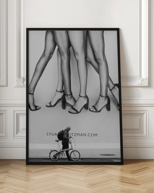 A black and white street photograph showing a stark contrast between a large fashion advertisement and a person on the sidewalk. The ad features multiple pairs of long female legs wearing high-heeled sandals. Below the ad, an older man with a backpack walks by, pushing a small foldable bicycle. Artwork
