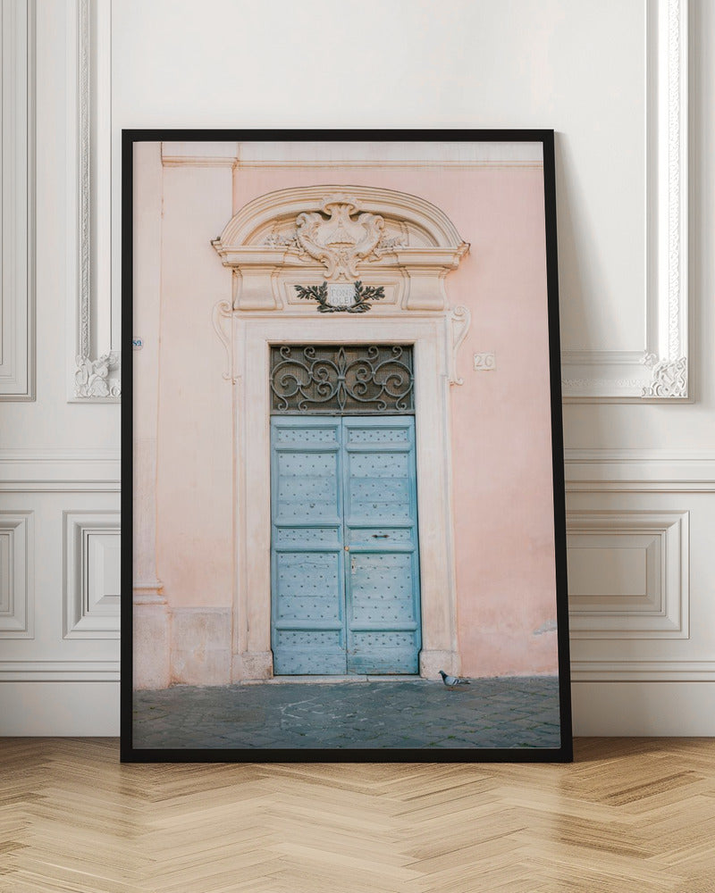 A light blue, studded double door set into a pale pink wall. The doorway is framed in white stone with an ornate pediment and a decorative wrought-iron transom window above the door. A single pigeon stands on the cobblestone pavement in front. Print