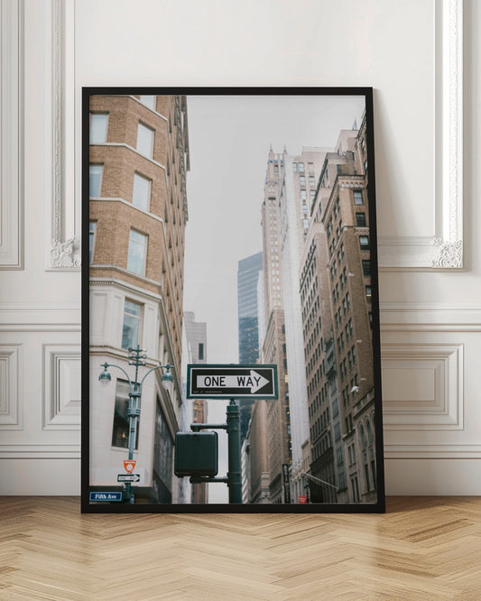 A framed photograph of a New York City street, with a prominent 'ONE WAY' sign in the foreground pointing right. The view is from a low angle, looking up between tall brick and stone buildings towards a bright, overcast sky. Wall Art