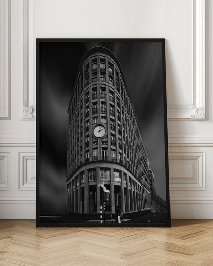 A dramatic low-angle, black and white photograph of a large, curved corner building with a prominent clock on its facade. The sky is dark with a motion blur effect, making the building appear grand and imposing. Artwork