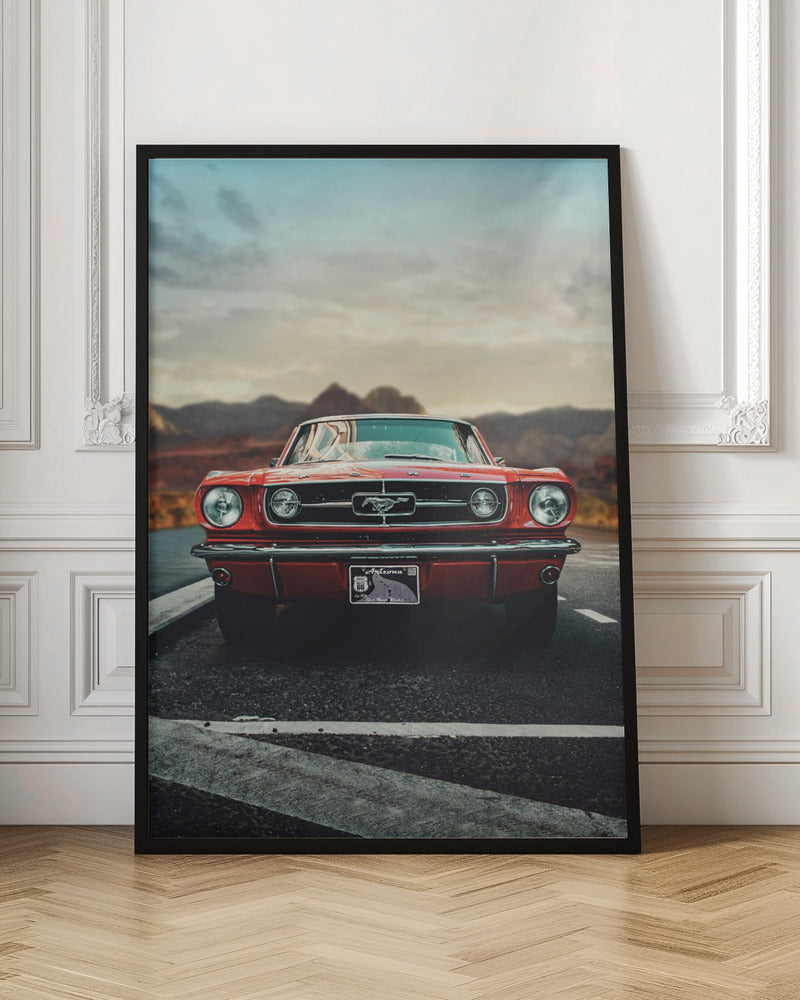 A low-angle, head-on shot of a vintage red Ford Mustang parked on an asphalt road. The iconic silver horse emblem is visible on the grille. In the background, a desert mountain range is blurred under a cloudy, muted sky. Poster