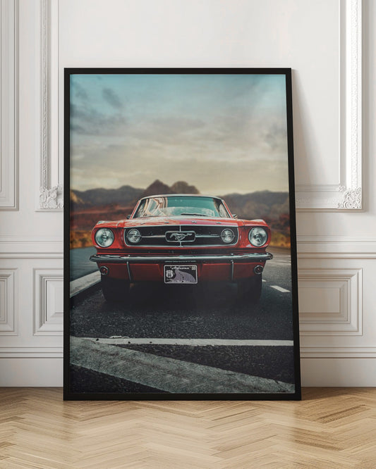 A low-angle, head-on shot of a vintage red Ford Mustang parked on an asphalt road. The iconic silver horse emblem is visible on the grille. In the background, a desert mountain range is blurred under a cloudy, muted sky. Poster