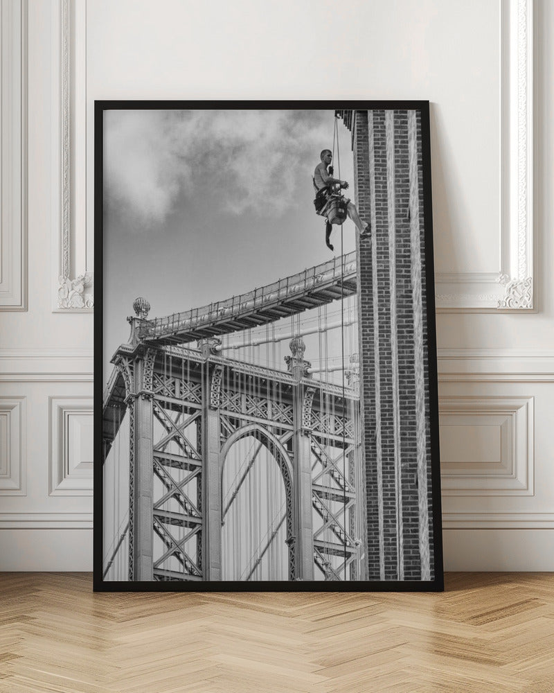 A black and white photograph capturing a window washer rappelling down the side of a tall brick building. In the background, the intricate steel structure of the Manhattan Bridge stands against a partly cloudy sky, creating a powerful urban scene. Decor