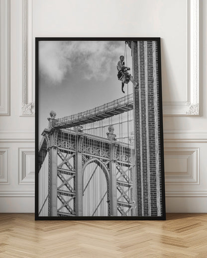 A black and white photograph capturing a window washer rappelling down the side of a tall brick building. In the background, the intricate steel structure of the Manhattan Bridge stands against a partly cloudy sky, creating a powerful urban scene. Decor