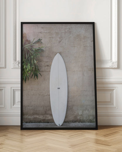 A minimalist photograph of a white surfboard leaning against a textured beige concrete wall. To the left, a large green staghorn fern adds a touch of nature. The entire scene is enclosed in a simple black frame. Artwork