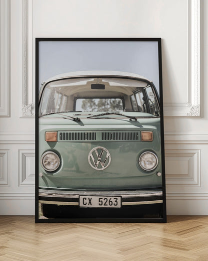 A close-up front view of a vintage sage green Volkswagen bus with a white roof. The iconic VW logo is on the front, with round headlights and a European license plate, set against a pale blue sky. Print