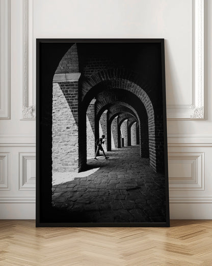 A dramatic black and white photograph of a person walking through a long brick arcade. Sunlight creates strong, contrasting shadows, highlighting the repeating arches that recede into the darkness, creating a powerful sense of depth and perspective. Decor