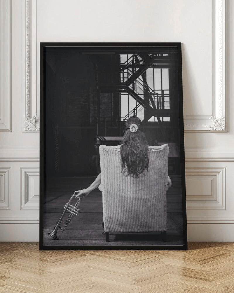 A black and white photograph of a woman seen from behind, sitting in a large, light-colored corduroy armchair. She has long, dark hair with a white flower pinned in it. Her left hand holds a trumpet, resting it on the dark floor of what appears to be an industrial warehouse with a metal staircase in the background. Wall Art
