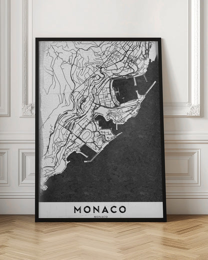 A modern black and white map of Monaco, presented as a framed poster. The map details the city's streets and coastline in black lines on a white background, while the sea is depicted in a solid, textured black. The title 'MONACO' is at the bottom in a bold, white band. Poster