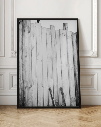 A black and white, low-angle photograph of a weathered wooden plank fence. The vertical planks are of varying heights, creating an uneven top line against a pale sky. The focus is on the texture of the wood, showing prominent grain, knots, and nail holes, all framed in black. Poster