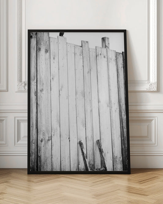A black and white, low-angle photograph of a weathered wooden plank fence. The vertical planks are of varying heights, creating an uneven top line against a pale sky. The focus is on the texture of the wood, showing prominent grain, knots, and nail holes, all framed in black. Poster