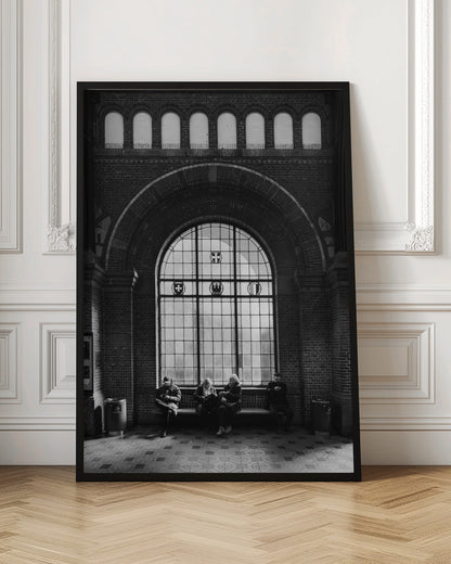 A dramatic black and white photograph of a large, brick-walled waiting area, possibly a train station. Four people are seated on a long bench beneath a massive arched window, which dominates the scene. The lighting creates a moody, contemplative atmosphere, highlighting the grandeur of the architecture. Artwork